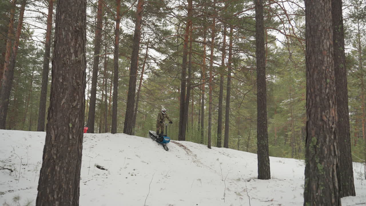 ATV rosso che si lancia in discesa attraverso una pineta innevata, il pilota perde il controllo e cade in un cumulo di neve, un quad blu scivola su un ripido pendio, segni di slittamento e rami sparsi, fredda luce di un cielo coperto.