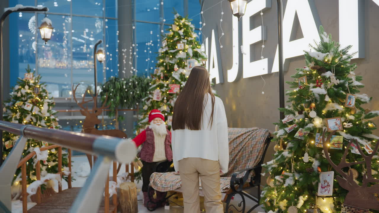 Lady walking up to decorative seats and sitting down in a Christmas-themed mall area with glowing inscription, surrounded by Christmas trees, lights, and festive decor