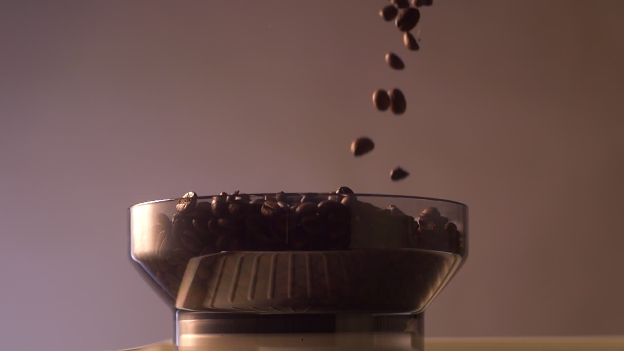 Chicchi di caffè cadono al rallentatore nella tramoggia di una macchina con portafiltro o di un macinacaffè per espresso. Il primo piano cinematografico cattura il flusso e il rimbalzo dei chicchi su uno sfondo con sfumatura delicata.