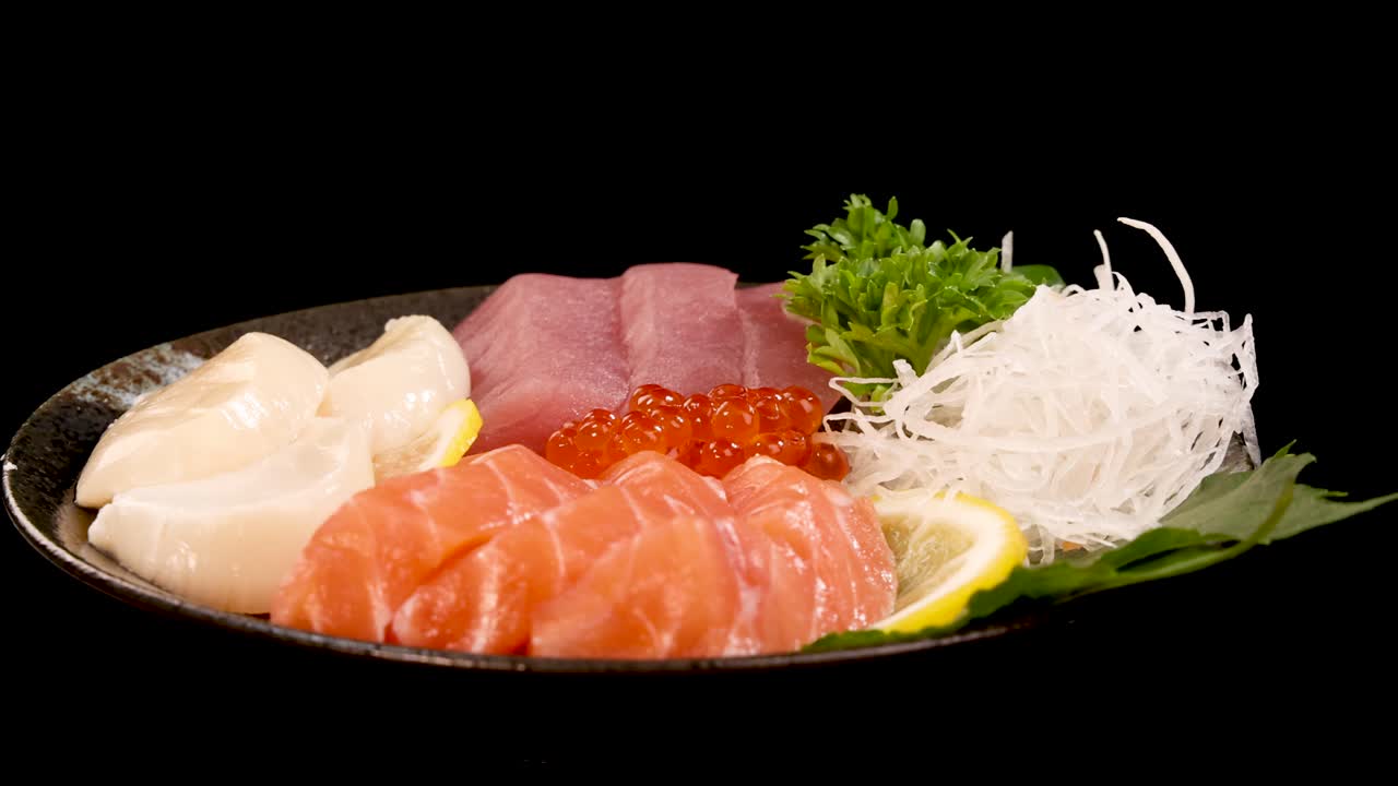 A plate of assorted sashimi, including salmon, tuna, scallop, and salmon roe, rotates smoothly under bright studio lighting against a seamless black backdrop