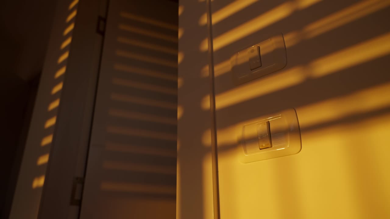 Electric or mechanical window blinds or shutter roll down at sunset or sunrise. Shadows appear on apartment or house wall with orange warm colors as sunlight shines through the cracks
