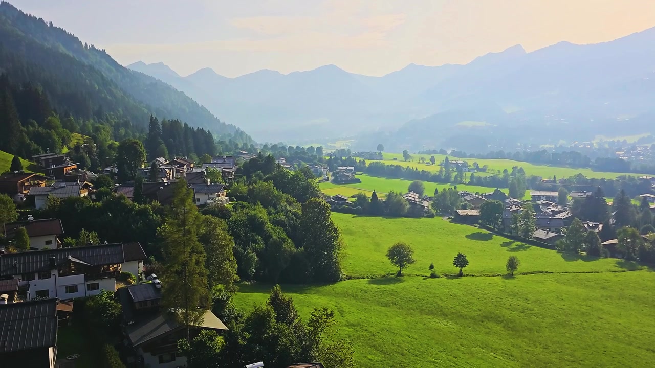 Salita in gondola sopra Kitzbühel, Austria, con tetti alpini, strade tortuose, colline lussureggianti, pendii boscosi e lontane montagne blu in un paesaggio vacanziero panoramico del Tirolo nelle Alpi