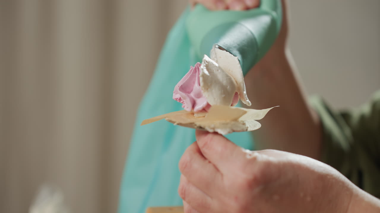 Close up of pink swirl candy delicately coated with icing using green piping bag held by baker, showing fine detail of dessert decoration process in warm kitchen environment