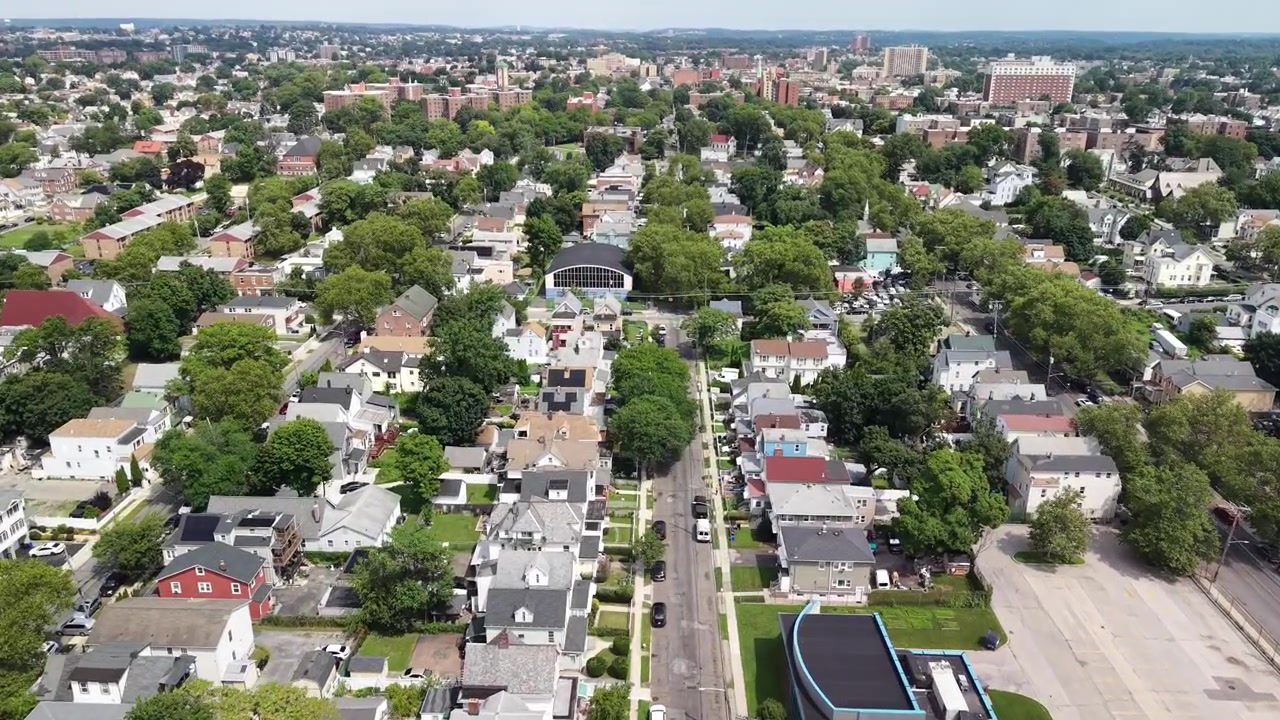 Vista aerea con ripresa all'indietro lungo una strada in un quartiere residenziale americano, che rivela lo skyline della città in lontananza in una giornata di sole con alberi verdi e case residenziali sottostanti