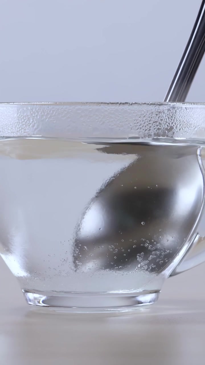 Un cucchiaio di metallo mescola polvere bianca in acqua calda all'interno di una tazza di vetro trasparente.