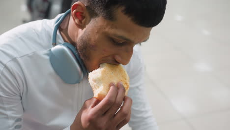 close up of boy seated on bench in gym holding sandwich with one hand preparing to take bite, wearing white top and black shorts, gym floor and equipment slightly blurred in background