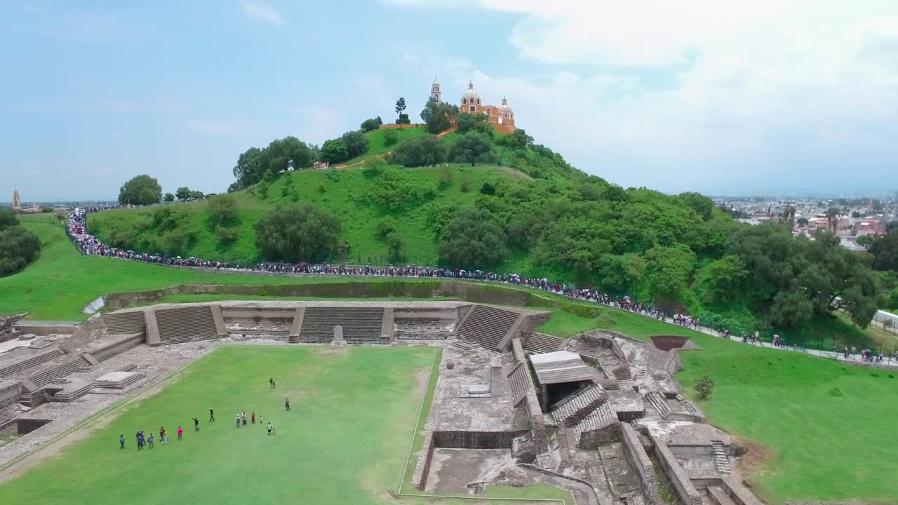 Ancient pyramid ruins and lush green hill with a church on top visited by a crowd of tourists
