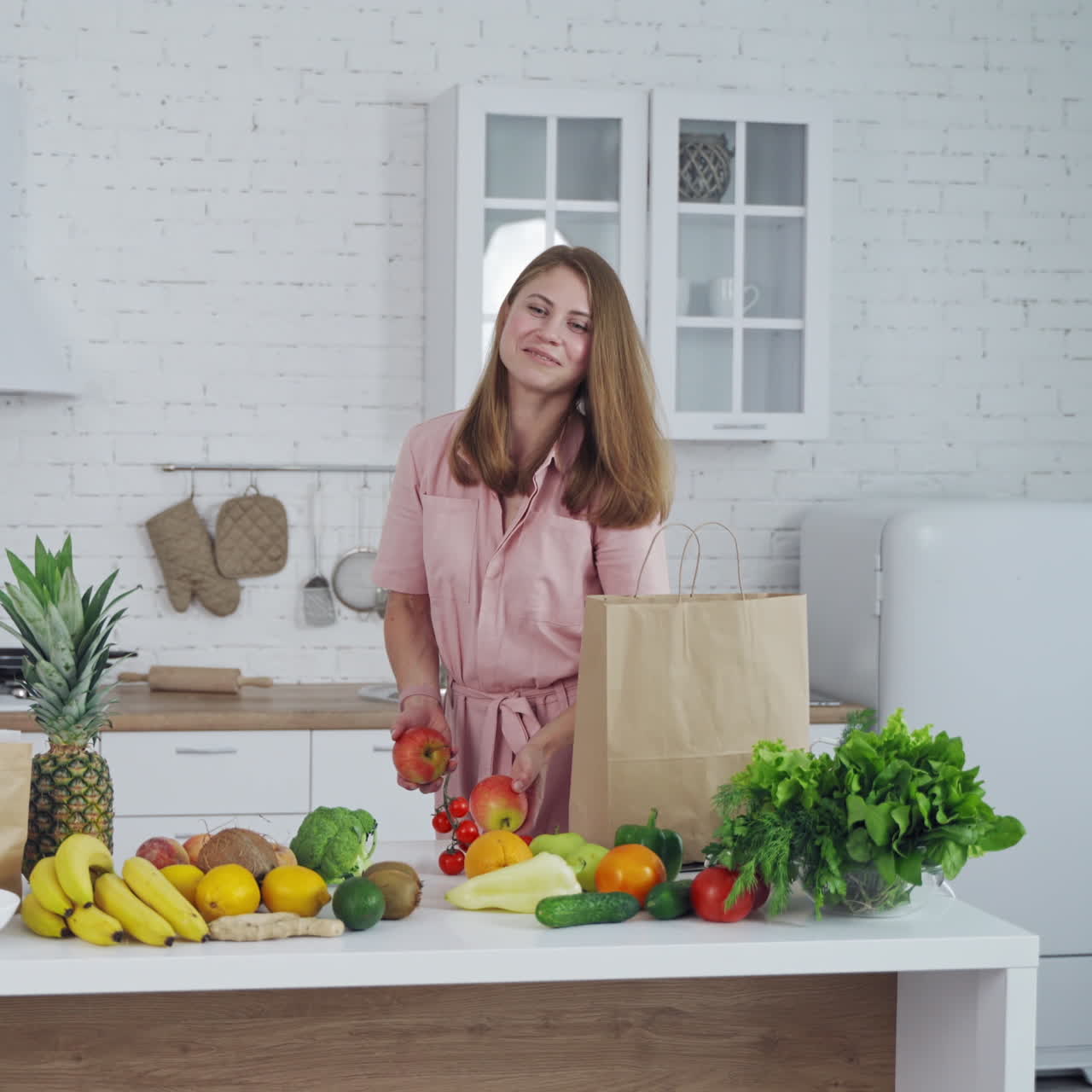 Happy young woman with fresh apples in the kitchen. Beautiful housewife holds and sniffs natural fruit on the background of healthy food on the table.