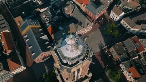 drone top-down view of birds flying in circles around a church tower's spire and cross in weert city center, limburg, during a clear morning