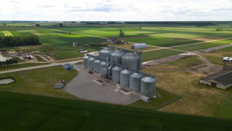 Vista-Aérea-De-Los-Silos-Agrícolas,-La-Planta-De-Fabricación-De-Graneros,-El-Almacenamiento-De-Productos-Agrícolas-Y-Los-Elevadores-De-Granos-En-Medio-De-Un-Campo-Cultivado-Verde,-Paralaje