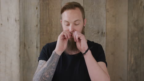 young hipster punk man touching beard looking pensive attractive caucasian male on wooden background