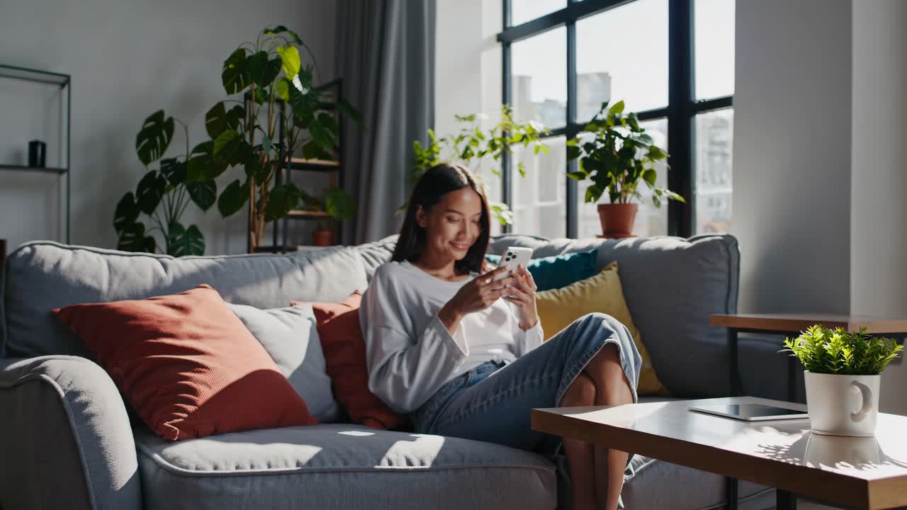 A young woman sits on a cozy sofa, smiling at her phone. The video captures a side angle