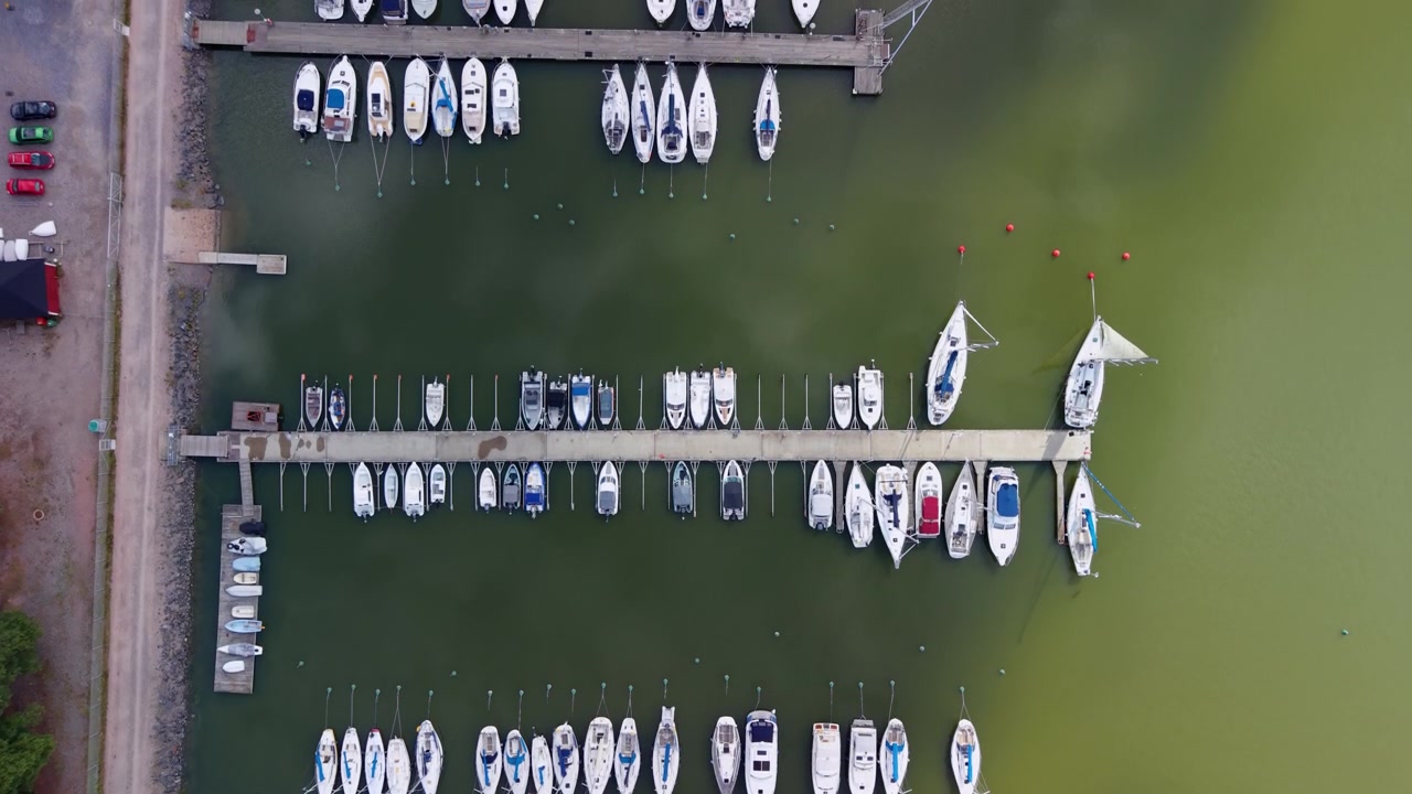 Ovenfra-udsigt over en marina i Helsinki, Finland, med rækker af sejlbåde og motorbåde fortøjet langs moler i roligt grønt vand ved siden af en kystvej.