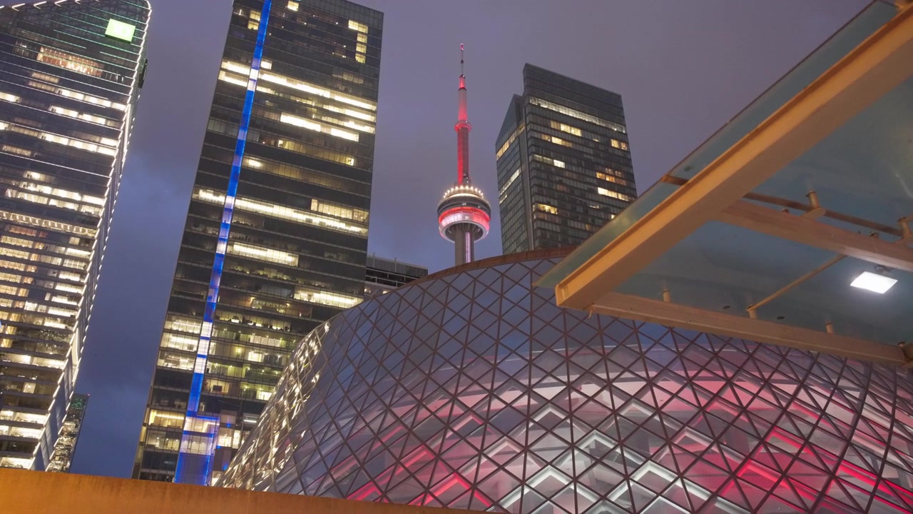 Lento movimento in avanti della telecamera verso la CN Tower incorniciata da torri per uffici illuminate e dal padiglione di vetro a Brookfield Place nel centro di Toronto di notte