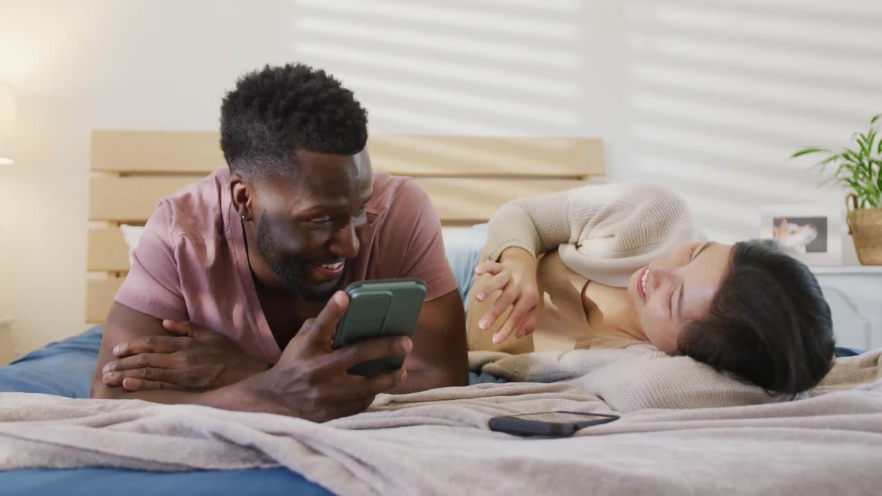 Felice coppia multietnica che usa lo smartphone e sdraiata in camera da letto