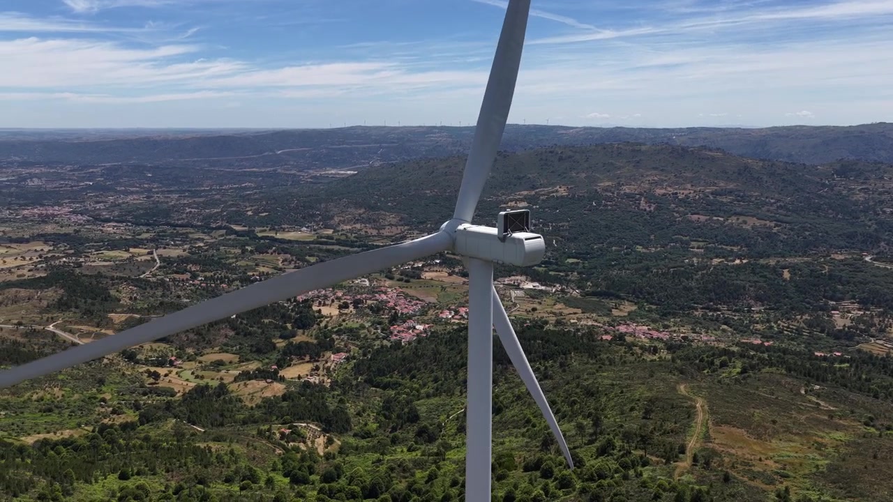 Vídeo impressionante de drone sobrevoa turbinas eólicas modernas girando sobre colinas verdes ondulantes, campos férteis e uma pitoresca aldeia portuguesa sob a luz solar intensa do meio-dia.