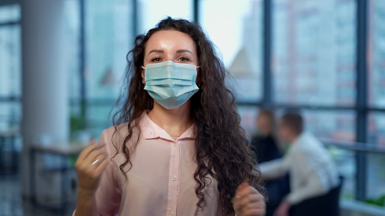 Dark-haired lady wearing mask standing in the middle of the office. Smiling female taking off mask gladly. Close up portrait.