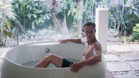 young sporty guy with muscular body rests in large bathtub at home