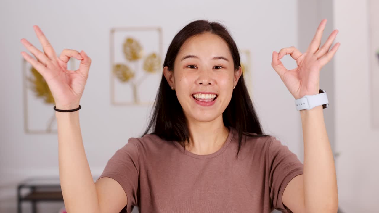 Young Asian woman smiles, makes OK hand gestures, well-lit modern home, static camera