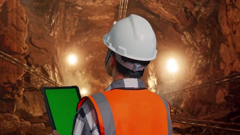 close up back view of asian male engineer with safety helmet working on a green screen tablet and looking around while standing in underground mine tunnel