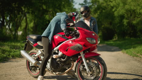 two sisters engage in conversation on a paved road surrounded by trees, one sister is on a red power bike, while the other, wearing a checkered shirt and black trousers, observes the bike