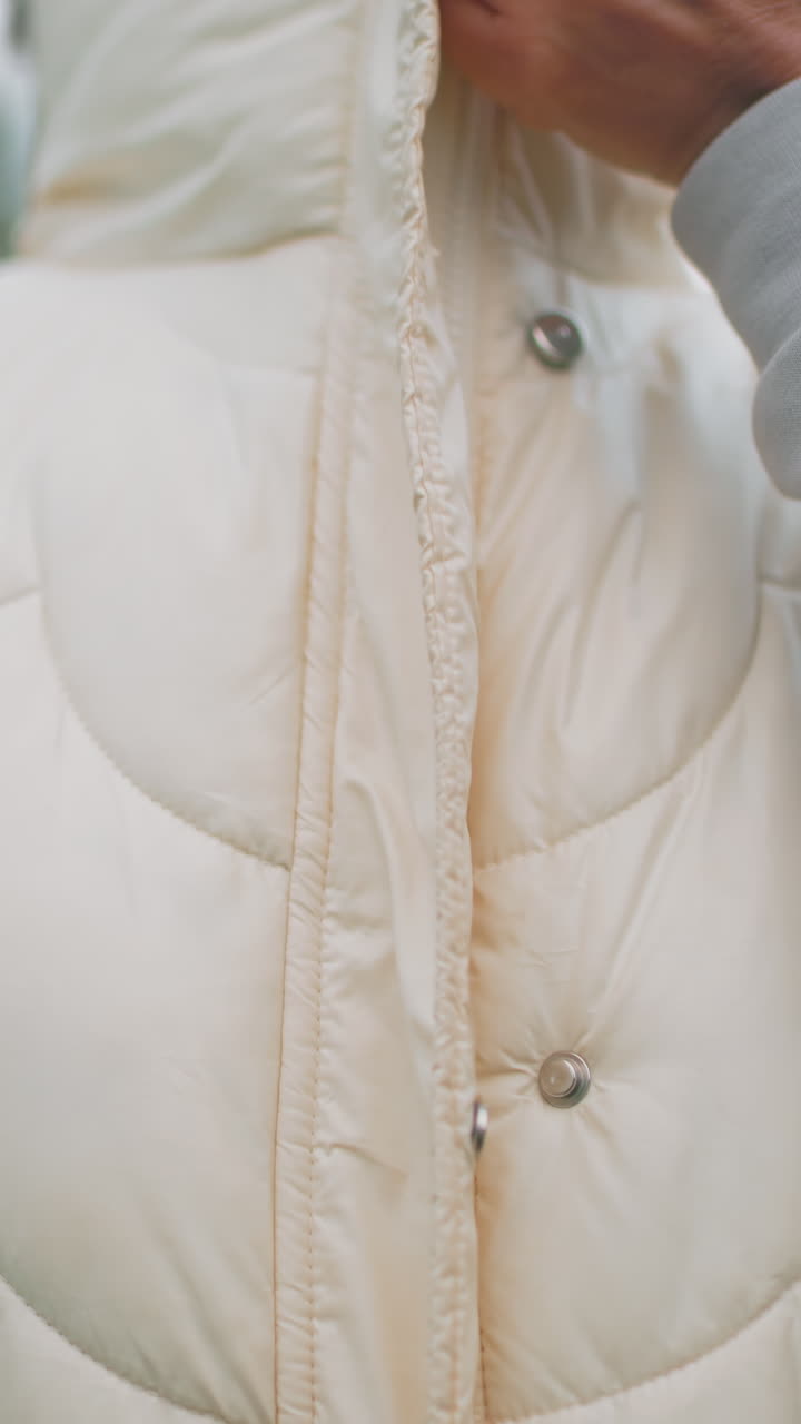 조끼 지퍼를 올리는 백인 여성의 클로즈업, 민트색 후드티가 안에 보임, 손으로 지퍼와 스냅을 조정, 부드러운 크림색 패딩 질감, 평온한 야외 아침, 부드러운 자기 관리 루틴, 평화로운 준비