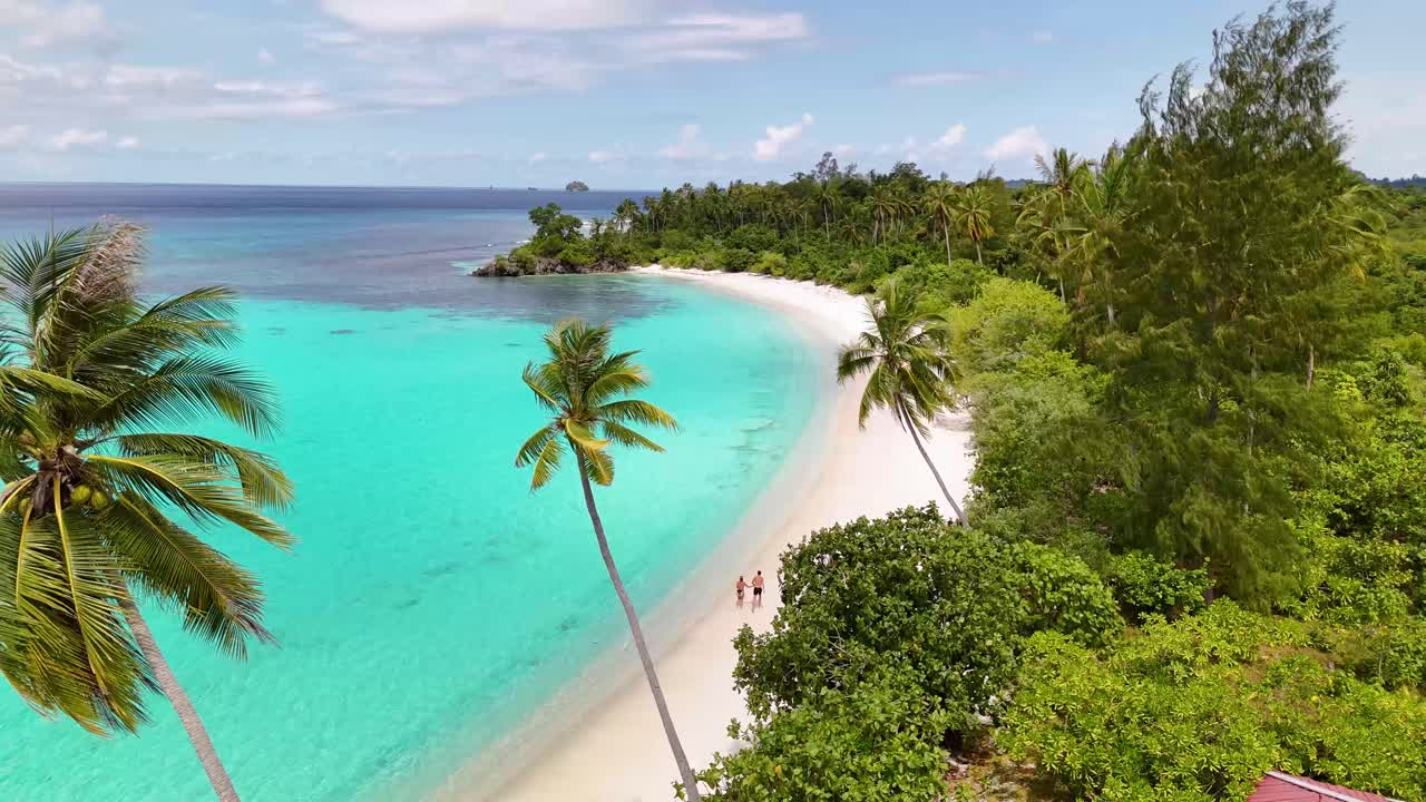 Video statico 4K da drone di una coppia che corre lungo la spiaggia di Oyama a Sulawesi, Indonesia. Una splendida spiaggia tropicale di sabbia bianca con un oceano blu brillante e bellissime palme verdi. Sembra il paradiso