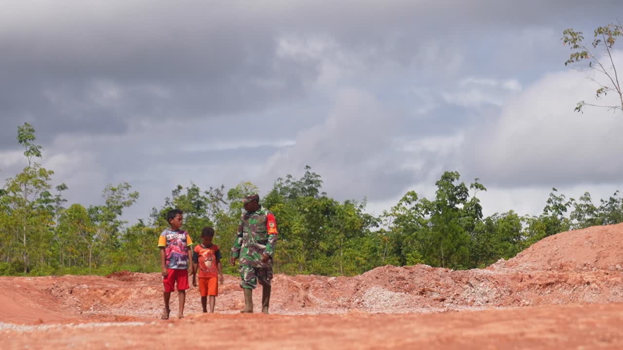 A soldier walks with two young boys on a dirt path in a rural landscape.