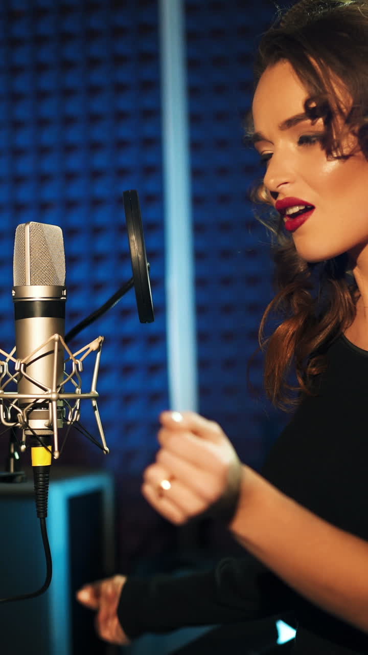 gorgeous woman with long hair is singing and dancing near the microphone in the audio studio on a blue background. Song recording. Vertical video