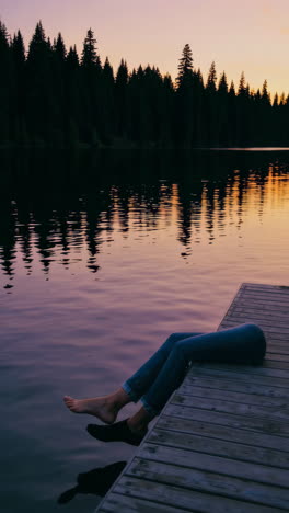 person dangling legs from a dock into a lake at sunset