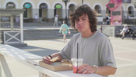 joven comiendo pizza y bebiendo una bebida fría solo mientras está de pie en una mesa al aire libre en la calle