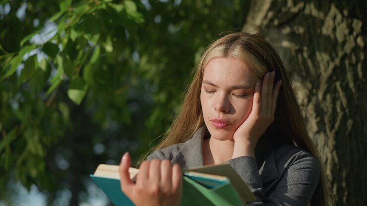 Lady in grey clothing sitting under a tree with her head resting on her right hand, holding an open book as sunlight softly illuminates her face, she appears deep in thought