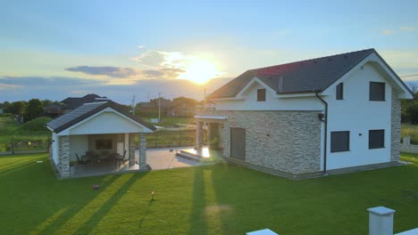 Aerial-drone-rotating-shot-of-two-story-brick-luxury-house-with-a-sitting-area-beside-the-house-surrounded-by-green-grass-garden-during-morning-time