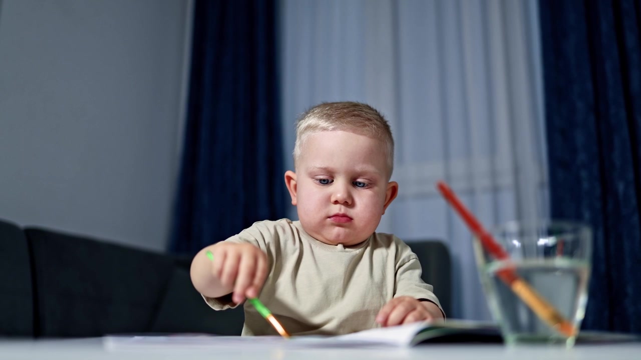 Bambino allegro di 2 anni è seduto al tavolo e disegna con i pennelli. Il bambino disegna con i colori e si diverte. Il bambino disegna e fa arte a casa