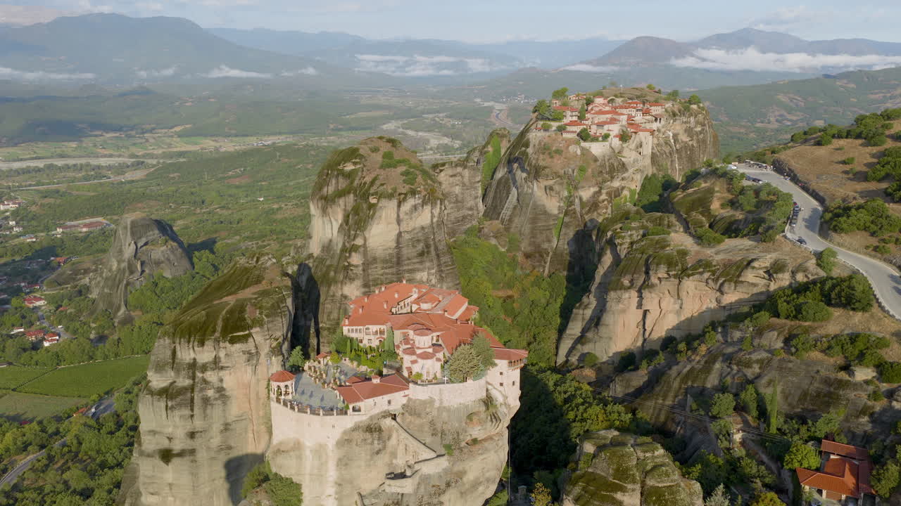 Cinematic aerial view of Meteora monastery in Greece perched on towering cliffs, dramatic rock formations and lush green valley create a breathtaking historic scene