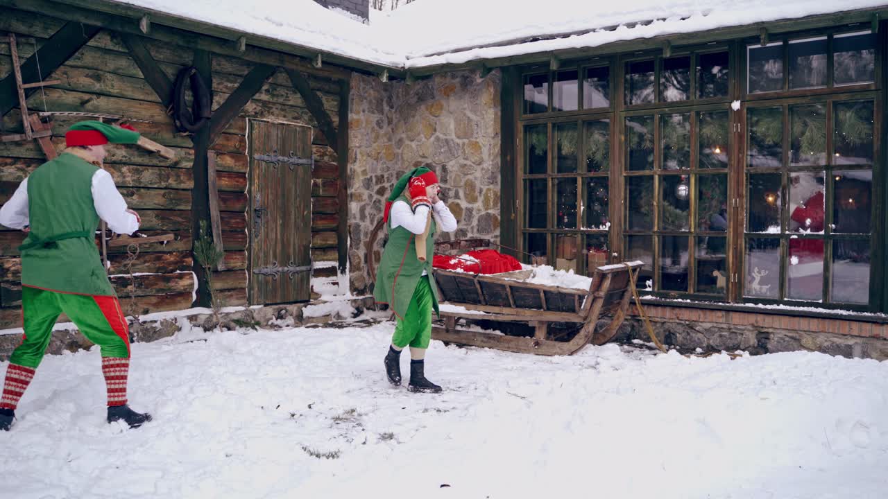 Two happy elves playing snowballs outdoors. Santa Claus looking through the window from the house. Christmas time.