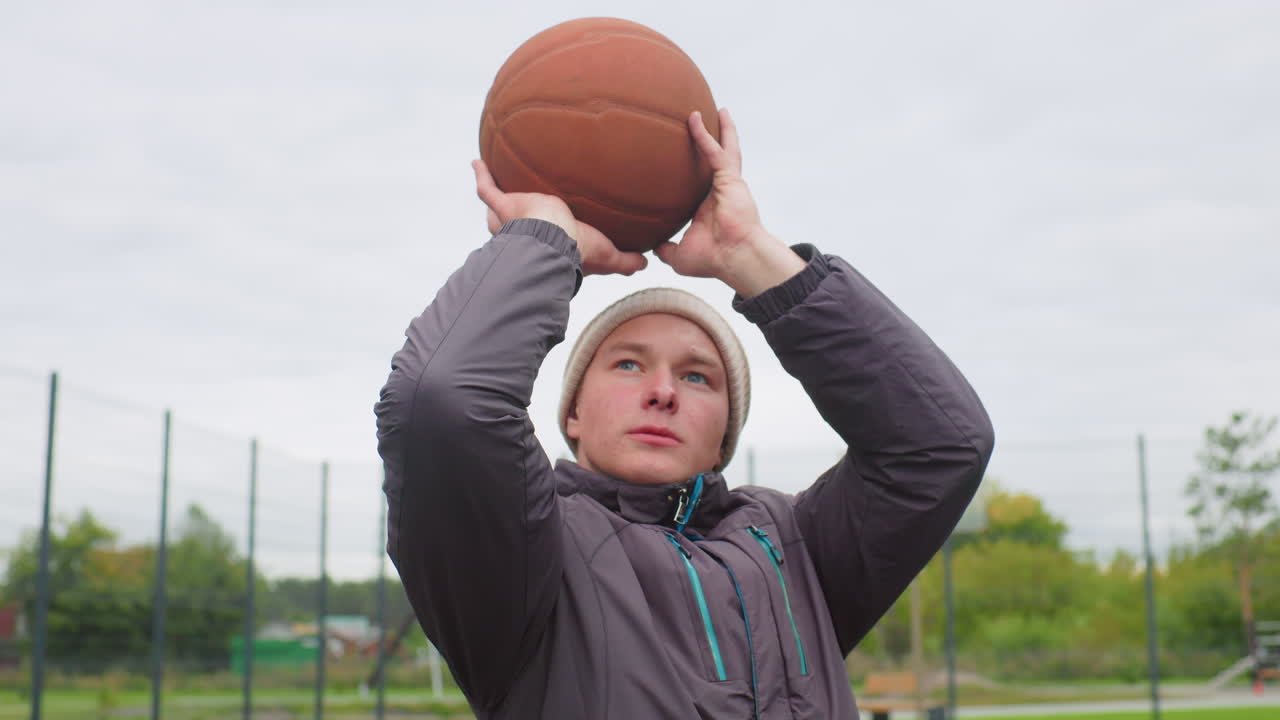 Ung atlet skyder basketball udendørs på indhegnet forstadbane iført hue og frakke, fokuseret udtryk, høj bue med rent efterløb, overskyet efterårshimmel, fjern legeplads og ensom