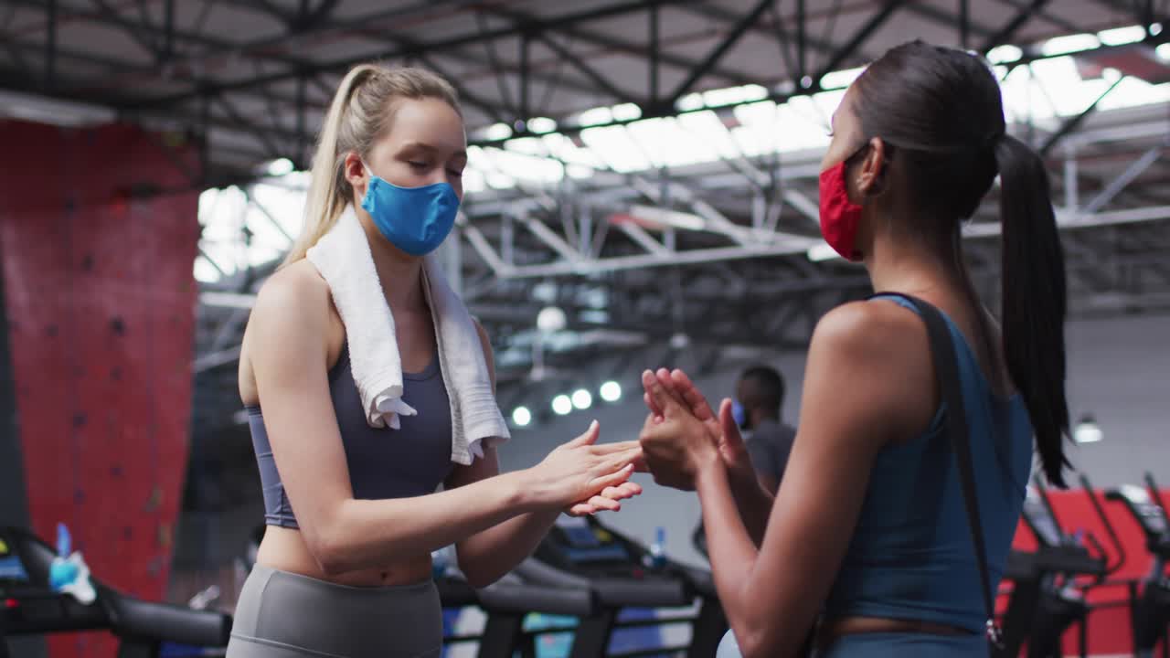 Two women at gym pressing dispenser, rubbing palms, animated graphics showing fitness safety