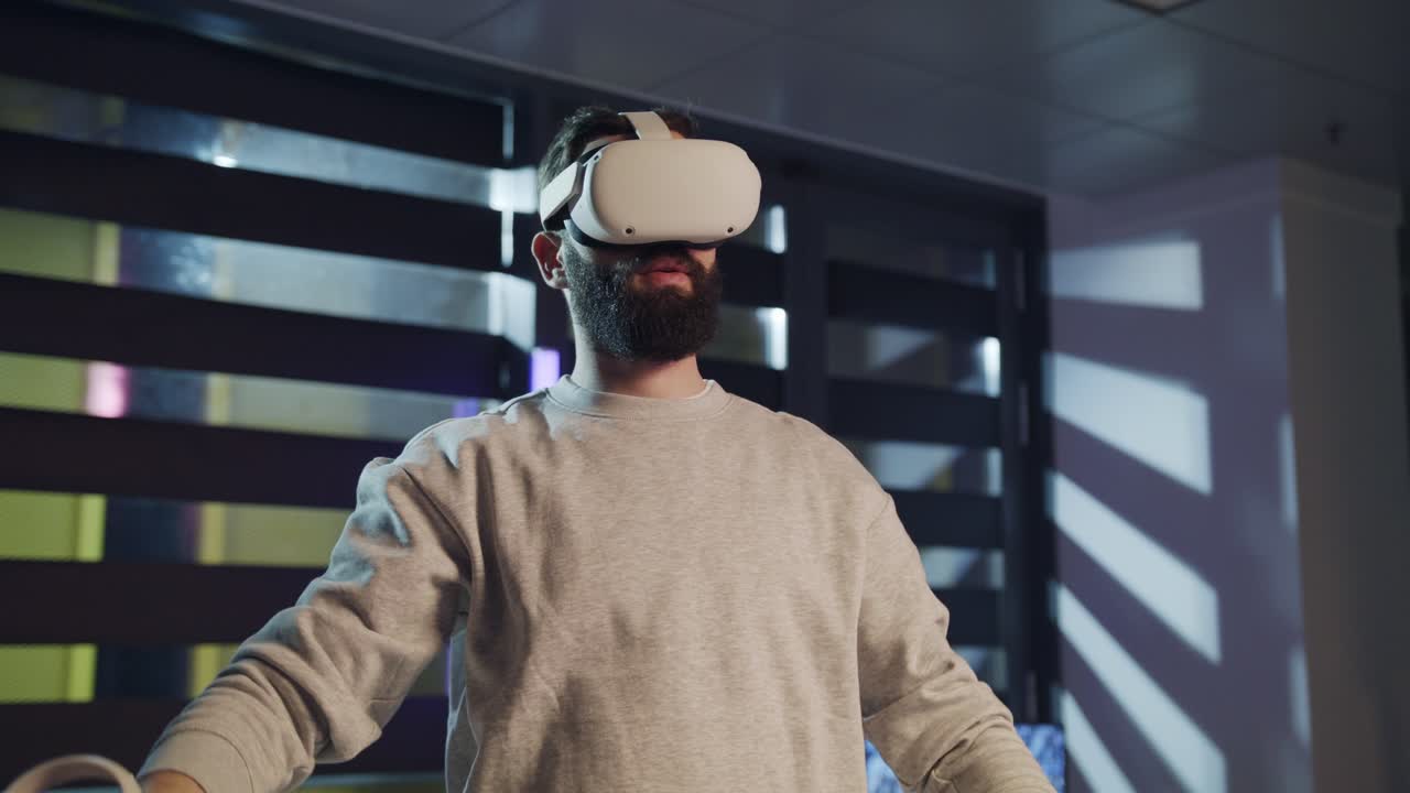 A bearded man wearing a VR headset lowers his controllers and stands still, fully immersed in a virtual experience. The room is lit by light coming through window blinds.
