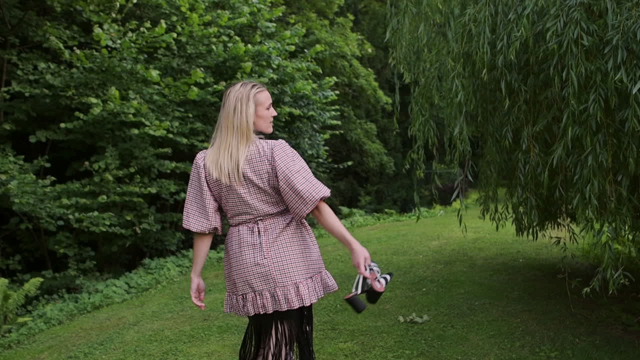 Fashion woman holding shoes in hand walking on grass through green public garden
