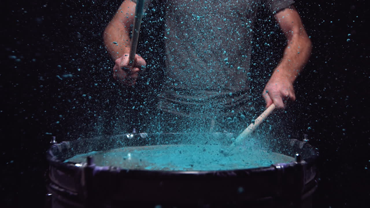 man die een trommel speelt die gevuld is met blauwe verf die spettert als de drumsticks in slow motion worden geraakt