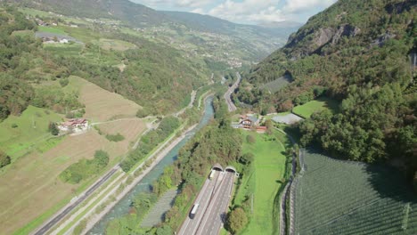 Highway-going-through-the-mountain-next-to-the-river,-cars-going-to-the-tunnel,-tilt-up-drone-view,-sunny-weather