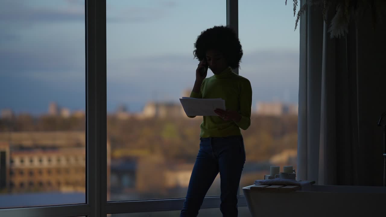 Silhouette of African American female manager talking on the phone holding documents standing at evening window indoors. Wide shot of young woman discussing project in home office.