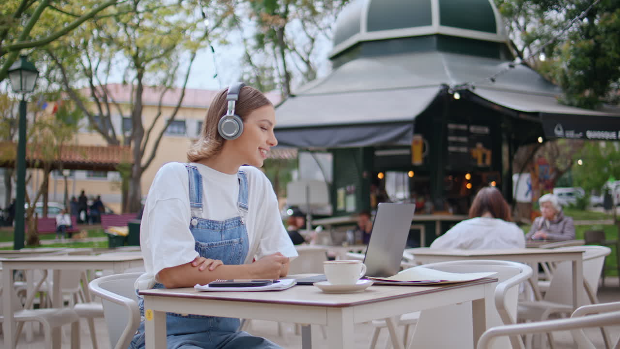 Cheerful girl greeting video call laptop at street cafe in headphones closeup