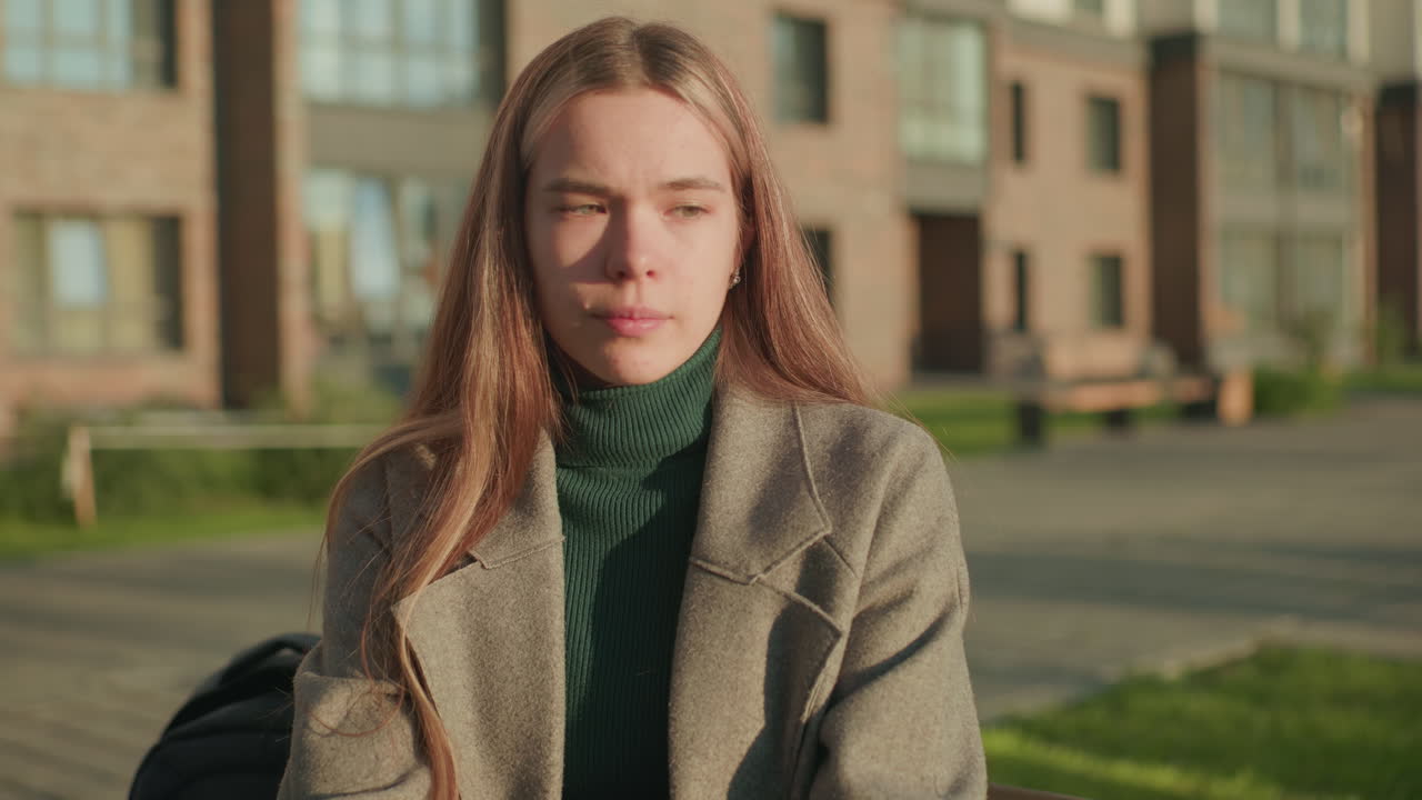 Young woman with long hair wearing green turtleneck and gray coat sits outdoors near residential buildings, eyes closed in calm expression while resting in natural sunlight