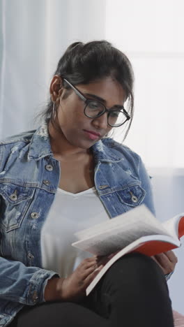 el lector indio enfocado da la vuelta a las páginas del libro sentado en la silla de la bolsa de frijoles en la sala de estar. la mujer con gafas disfruta leyendo en casa contra una ventana brillante