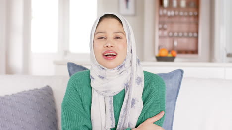 happy biracial woman in hijab waving to camera at home, copy space, slow motion