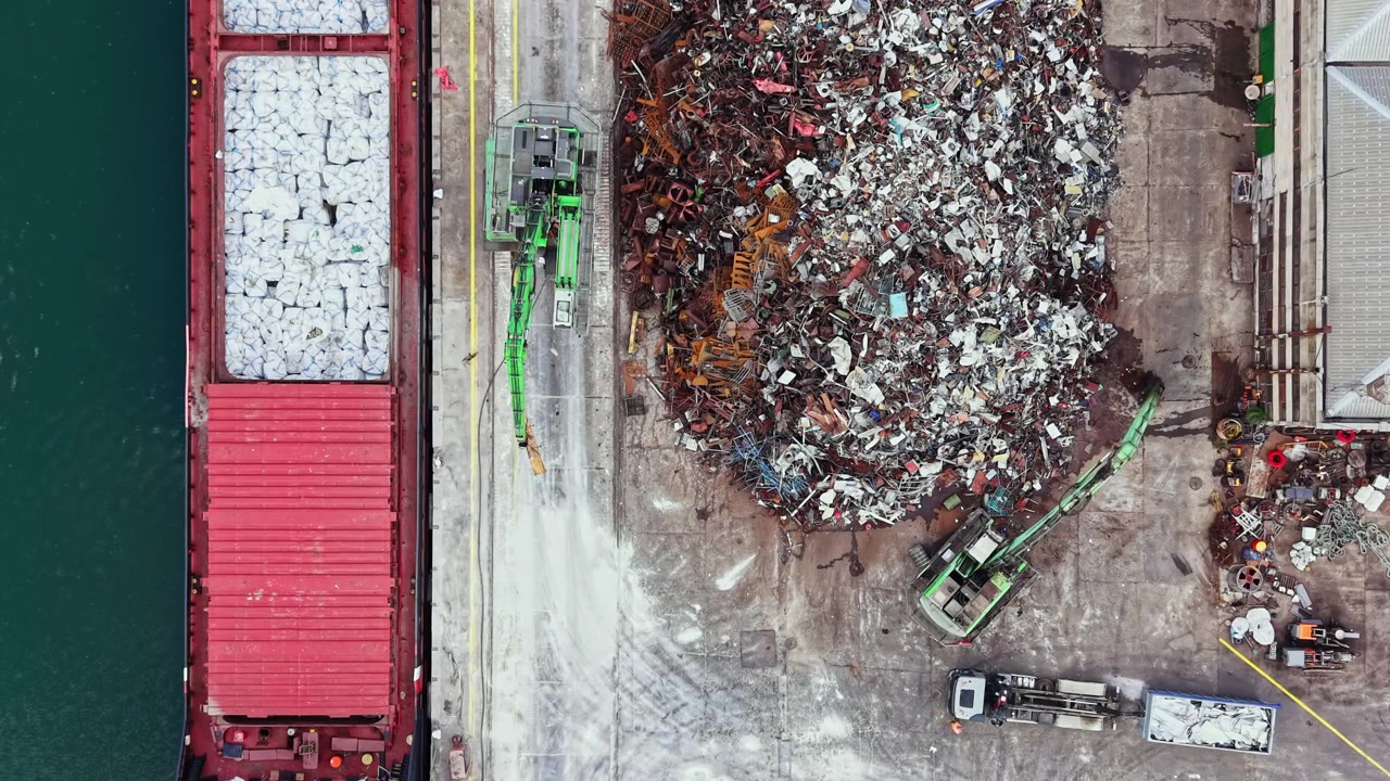 Large cargo ship docks at a recycling facility, where heavy machinery sorts and moves piles of metal and other scrap materials for processing during the day