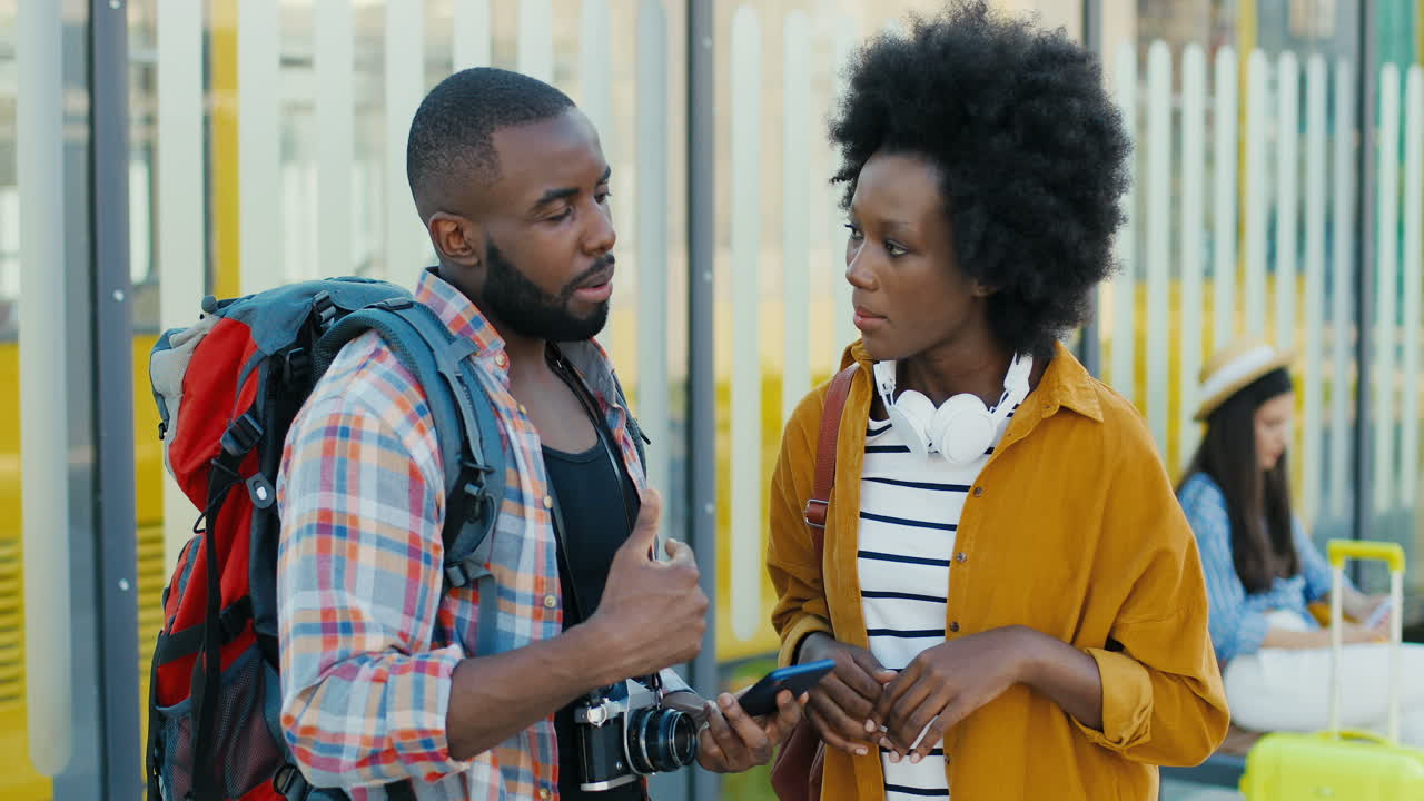jóvenes afroamericanos felices hombres y mujeres viajeros en la parada de autobús hablando y usando un teléfono inteligente para planificar una ruta