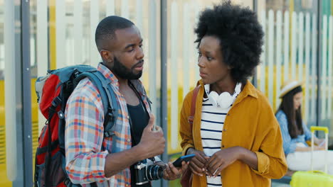 african american young happy man and woman travellers at bus stop talking and using smartphone to plan a route